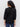 A woman with curly hair stands with her back to the camera, wearing Sanctuary Clothing’s black petal drop blouse and blue jeans against a plain light background.