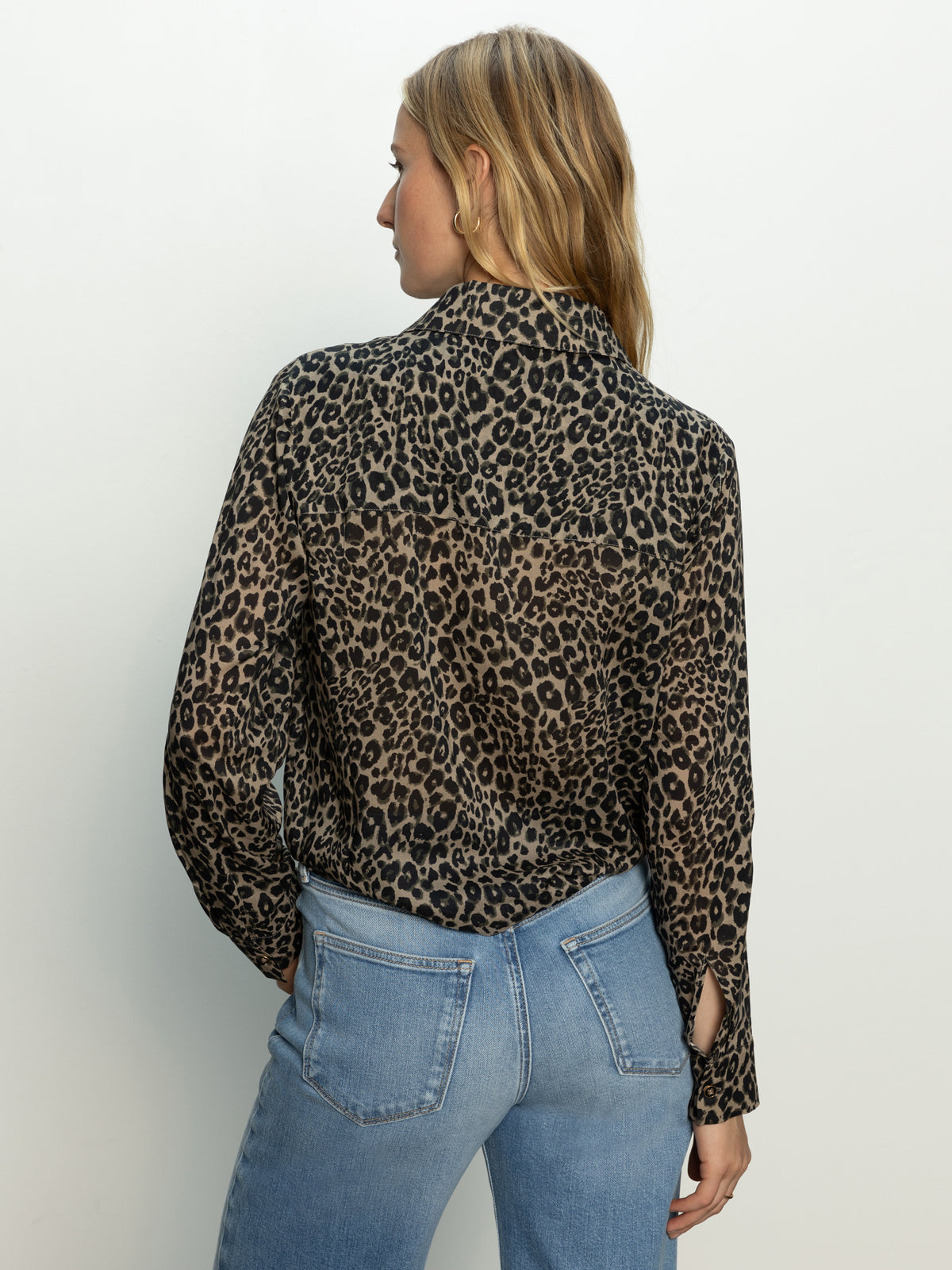 A woman with blonde hair faces away, wearing the Sanctuary Clothing wild love tie top french kitty— a long-sleeve leopard print blouse— and light blue jeans against a plain light background.