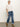 A woman with blonde hair tied back and a neutral expression poses against a plain background in Sanctuary Clothing's Deja Vu Blouse Light Oat—sheer with ruffled sleeves—paired with blue cuffed jeans and tan suede ankle boots.
