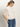 A blonde woman with a ponytail stands sideways against a plain background, wearing Sanctuary Clothing's Deja Vu Blouse in Light Oat, a sheer white short-sleeve top with ruffled details, paired with blue jeans.