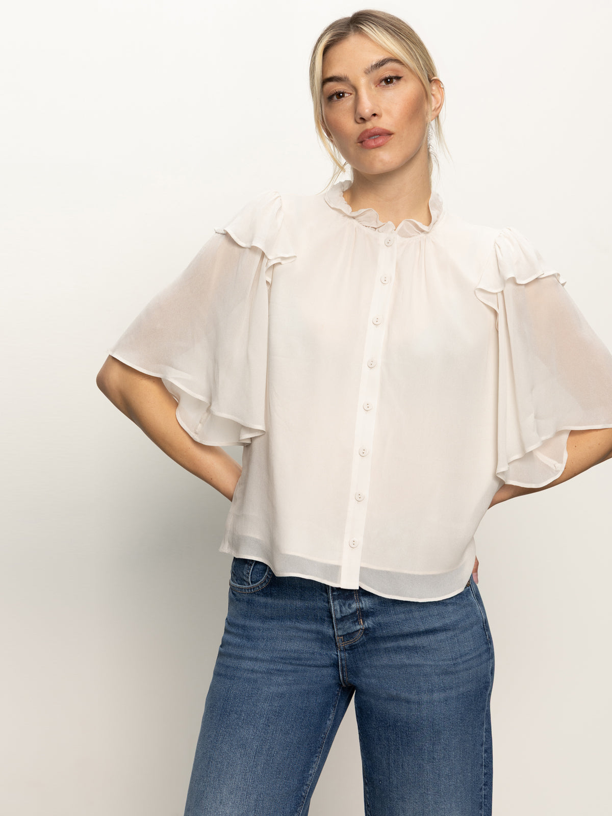 A woman with light hair tied back stands against a plain background wearing the Sanctuary Clothing Deja Vu Blouse in Light Oat, a sheer, short-sleeved top with ruffle details, paired with blue jeans and a neutral expression, hands on her hips.