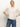 A woman with light hair tied back stands against a plain background wearing the Sanctuary Clothing Deja Vu Blouse in Light Oat, a sheer, short-sleeved top with ruffle details, paired with blue jeans and a neutral expression, hands on her hips.