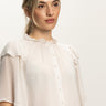 A woman with blonde hair in a ponytail wears the Sanctuary Clothing Deja Vu Blouse in Light Oat, featuring ruffled sleeves and front buttons, standing against a plain white background.