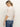A woman with blonde hair in a ponytail wears the Sanctuary Clothing Deja Vu Blouse in Light Oat and blue jeans, standing against an off-white background, turned slightly away from the camera.