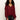 Woman with long curly hair wears a loose, sheer burgundy blouse with buttons and ruffled sleeves, paired with black leather pants, standing against a plain light background.