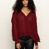 Woman with long curly hair wears a loose, sheer burgundy blouse with buttons and ruffled sleeves, paired with black leather pants, standing against a plain light background.
