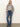 A woman with straight blonde hair and a neutral expression stands against a plain wall wearing the Sanctuary Clothing wild dreamer blouse mother leo, high-waisted blue jeans, a black belt, and black heeled boots.