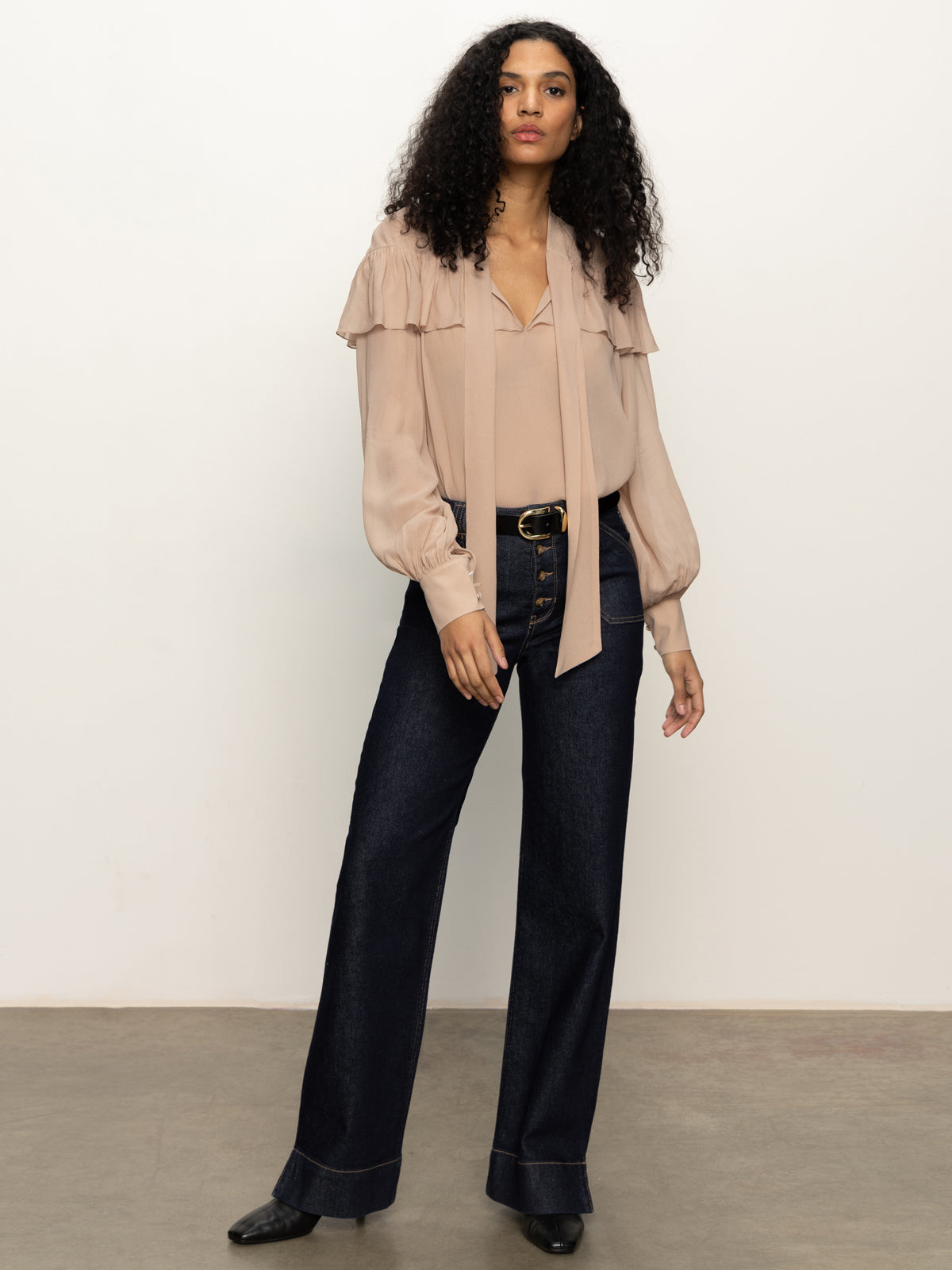 A woman with curly hair models the Sanctuary Clothing "fade into you ruffle blouse" in fawn, paired with high-waisted dark blue wide-leg jeans and black shoes, standing against a plain light background.