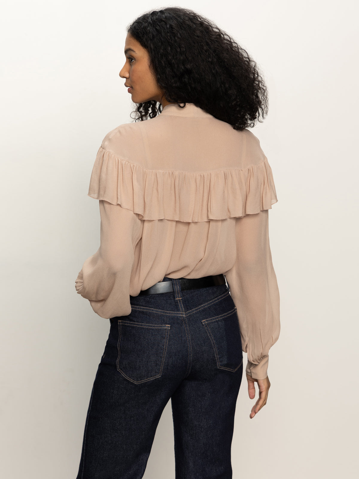 A woman with curly dark hair, wearing the Sanctuary Clothing "fade into you ruffle blouse" in fawn and dark blue jeans, stands facing away from the camera against a plain light background.