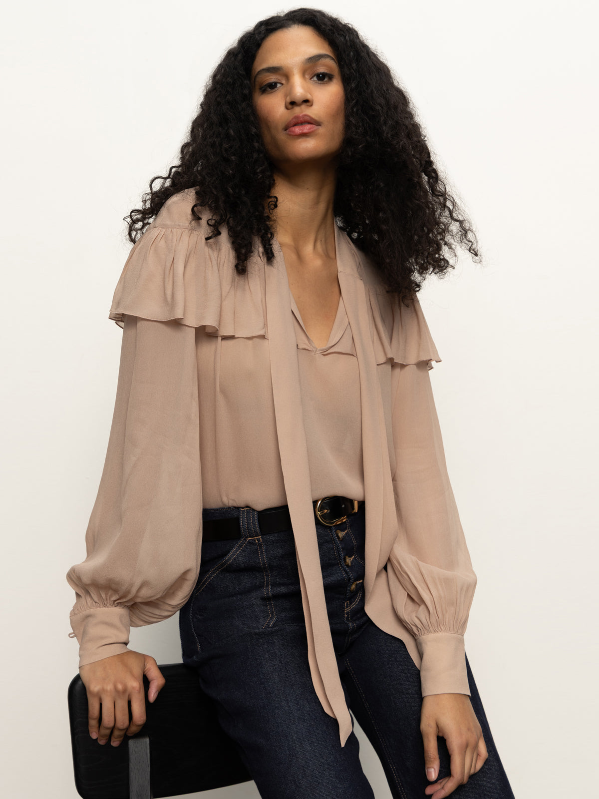 A woman with long curly hair sits on a black stool, wearing the fade into you ruffle blouse in fawn by Sanctuary Clothing, styled with dark high-waisted jeans and a black belt, against a plain light background.