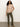 A woman with curly hair wears the Sanctuary Clothing sweet romance lace top fawn, styled with olive green pants and tan heeled boots, standing against a plain light background with one hand in her pocket.