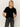 A woman with long blonde hair wears the Sanctuary Clothing "we belong together" velvet top in black, paired with dark gray jeans and a black belt, standing against a plain light background with her hands in her pockets.