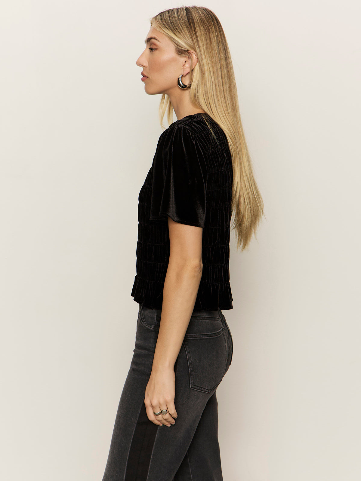 A woman with long blonde hair stands in profile against a plain light background, wearing the Sanctuary Clothing "we belong together" black velvet top and dark high-waisted jeans.