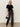 A woman with long blonde hair and gold hoop earrings stands against a plain background wearing the Sanctuary Clothing "we belong together velvet top" in black, paired with dark wide-leg jeans, black boots, and a studded black handbag.