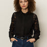 A woman with curly brown hair stands with her hands in her pockets against a light background, wearing the Sanctuary Clothing serendipity lace tuxedo shirt in black and blue jeans.