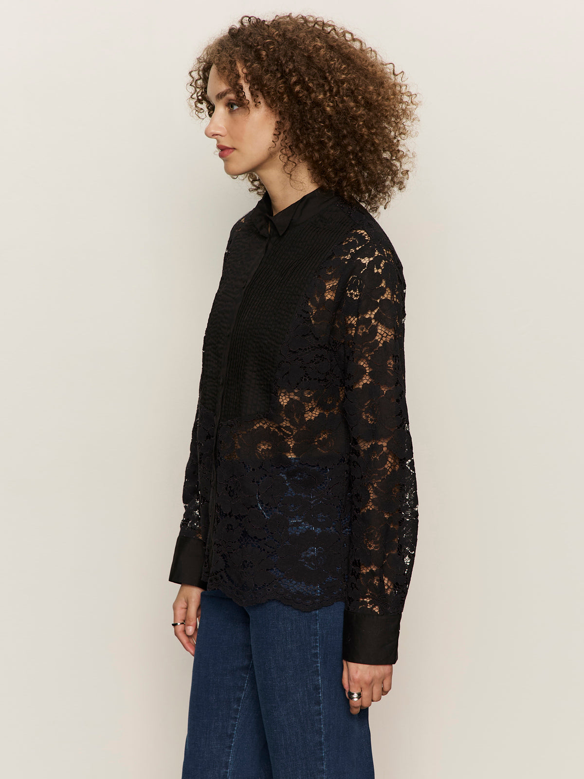 A woman with curly hair stands in profile against a light backdrop, wearing the Sanctuary Clothing Serendipity Lace Tuxedo Shirt in black over a black top and blue jeans.
