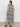 A person stands with their back to the camera in a Sanctuary Clothing contrast trim maxi dress featuring black macrame stripes and side slits. They pair this sleeveless look with black sandals against a plain white wall.