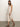 A woman with brown hair stands on a concrete floor wearing the Sanctuary Clothing contrast trim maxi dress in sandstone white stripe, featuring a sleeveless, knee-length design with a side slit. She pairs it with brown sandals against a plain white background.