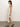 A woman with long dark hair wears a Sanctuary Clothing "contrast trim maxi dress" featuring white and beige horizontal stripes. She stands sideways, facing left, with her right leg forward, paired with brown sandals against a plain backdrop.