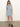 A woman stands indoors against a white wall and concrete floor, wearing Sanctuary Clothing’s ila denim dress bouquet—a sleeveless light blue button-front style with a matching waist tie—paired with black strappy sandals.
