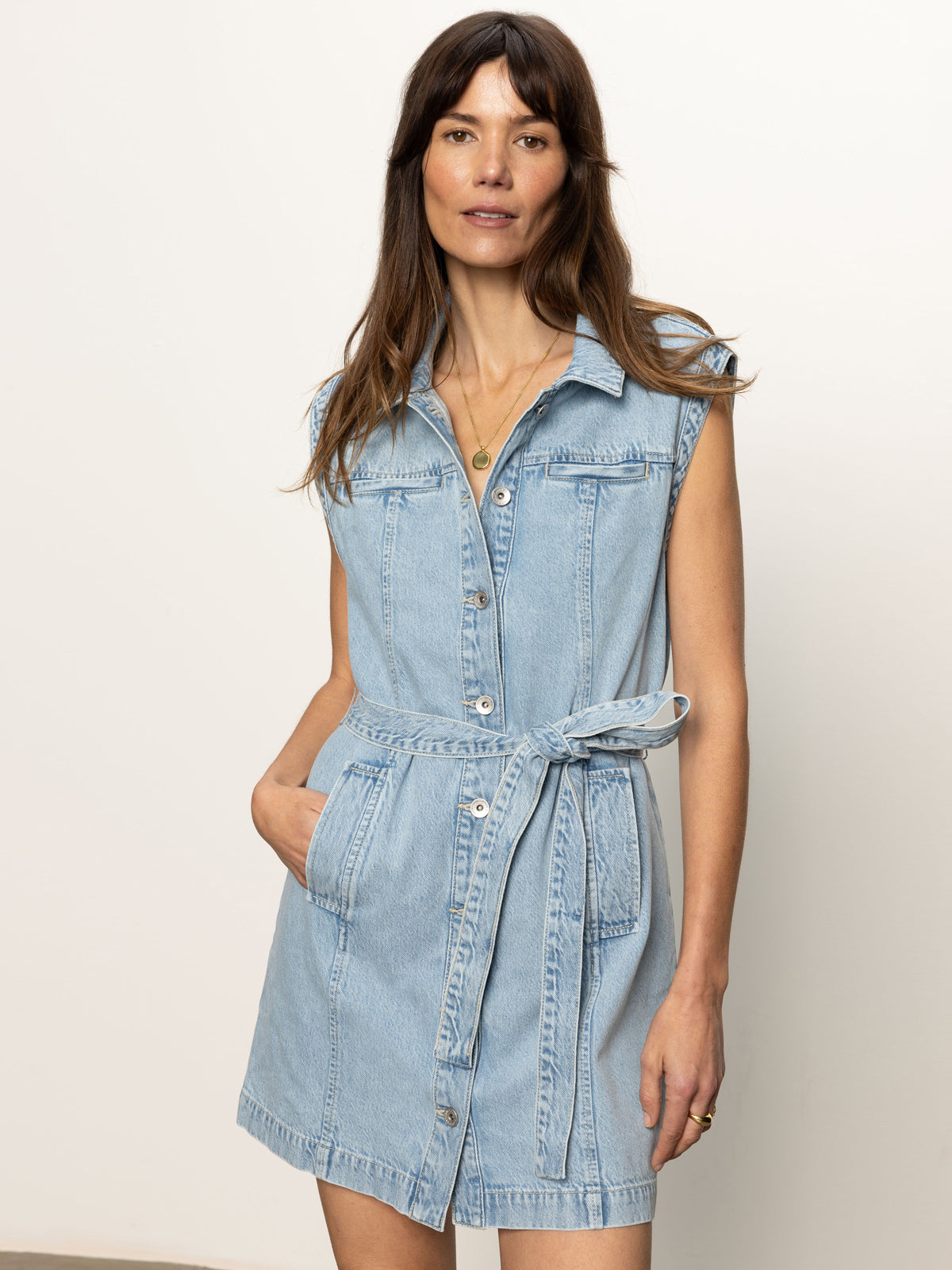A woman with long brown hair models the Sanctuary Clothing ila denim dress bouquet—a sleeveless, light blue denim dress with a tie belt and front buttons—standing against a plain background with her hand in one pocket.