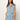 A woman with long brown hair models the Sanctuary Clothing ila denim dress bouquet—a sleeveless, light blue denim dress with a tie belt and front buttons—standing against a plain background with her hand in one pocket.