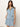 A woman with long brown hair models the Sanctuary Clothing ila denim dress bouquet”a sleeveless, light blue denim dress with a tie belt and front buttons”standing against a plain background with her hand in one pocket.