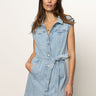 A woman with long brown hair models the Sanctuary Clothing ila denim dress bouquet—a sleeveless, light blue denim dress with a tie belt and front buttons—standing against a plain background with her hand in one pocket.