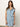A woman with long brown hair models the Sanctuary Clothing "ila denim dress bouquet," a sleeveless light blue button-front dress with a waist tie, standing against a plain light background.