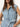 A woman wears the Sanctuary Clothing ila denim dress bouquet, a sleeveless light blue button-up style with a tied waist belt and pockets, standing against a plain white background.