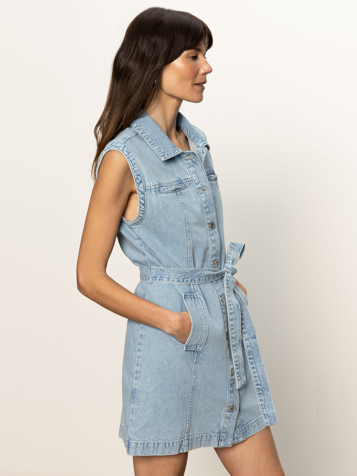 A woman stands sideways against a plain light background wearing the Sanctuary Clothing ila denim dress bouquet, a sleeveless, light blue denim dress with a collar, button-down front, waist tie, and pockets.
