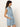 A woman stands sideways against a plain light background wearing the Sanctuary Clothing ila denim dress bouquet, a sleeveless, light blue denim dress with a collar, button-down front, waist tie, and pockets.