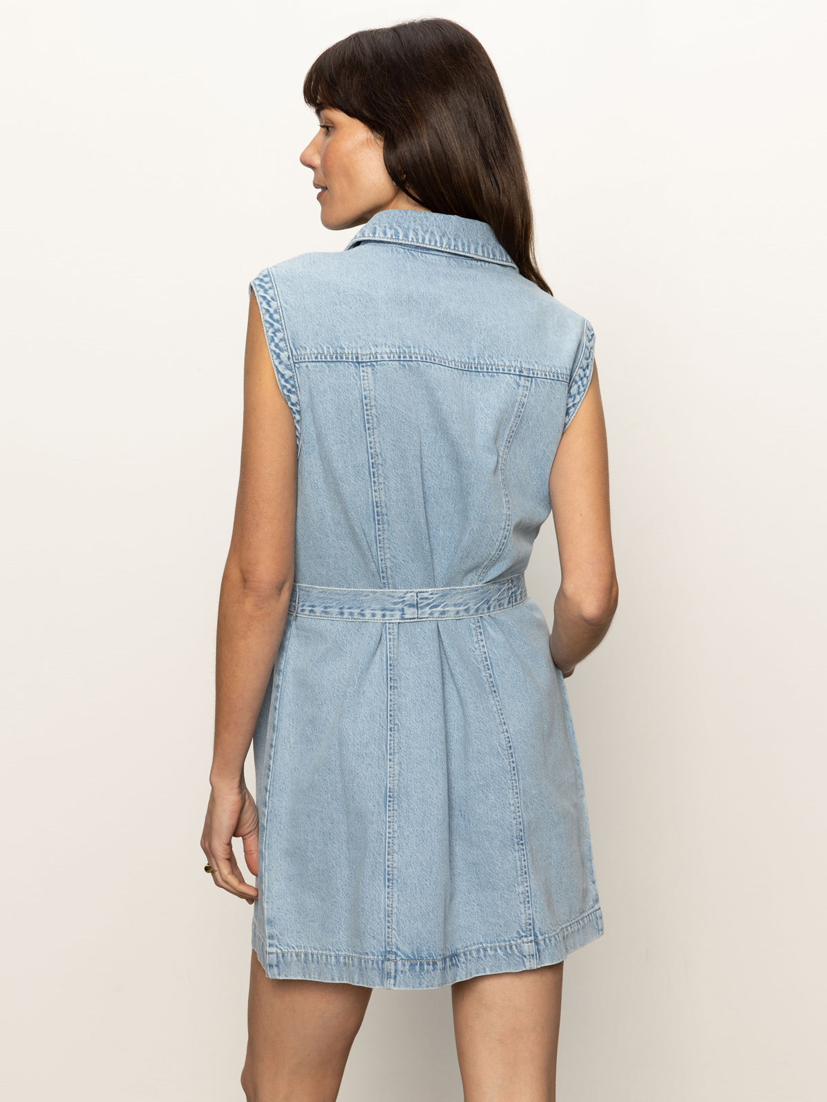 A woman with long brown hair, seen from the back, wears the Sanctuary Clothing ila denim dress bouquet—a sleeveless light blue belted denim dress—standing against a plain light background.
