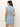 A woman with long brown hair, seen from the back, wears the Sanctuary Clothing ila denim dress bouquet—a sleeveless light blue belted denim dress—standing against a plain light background.