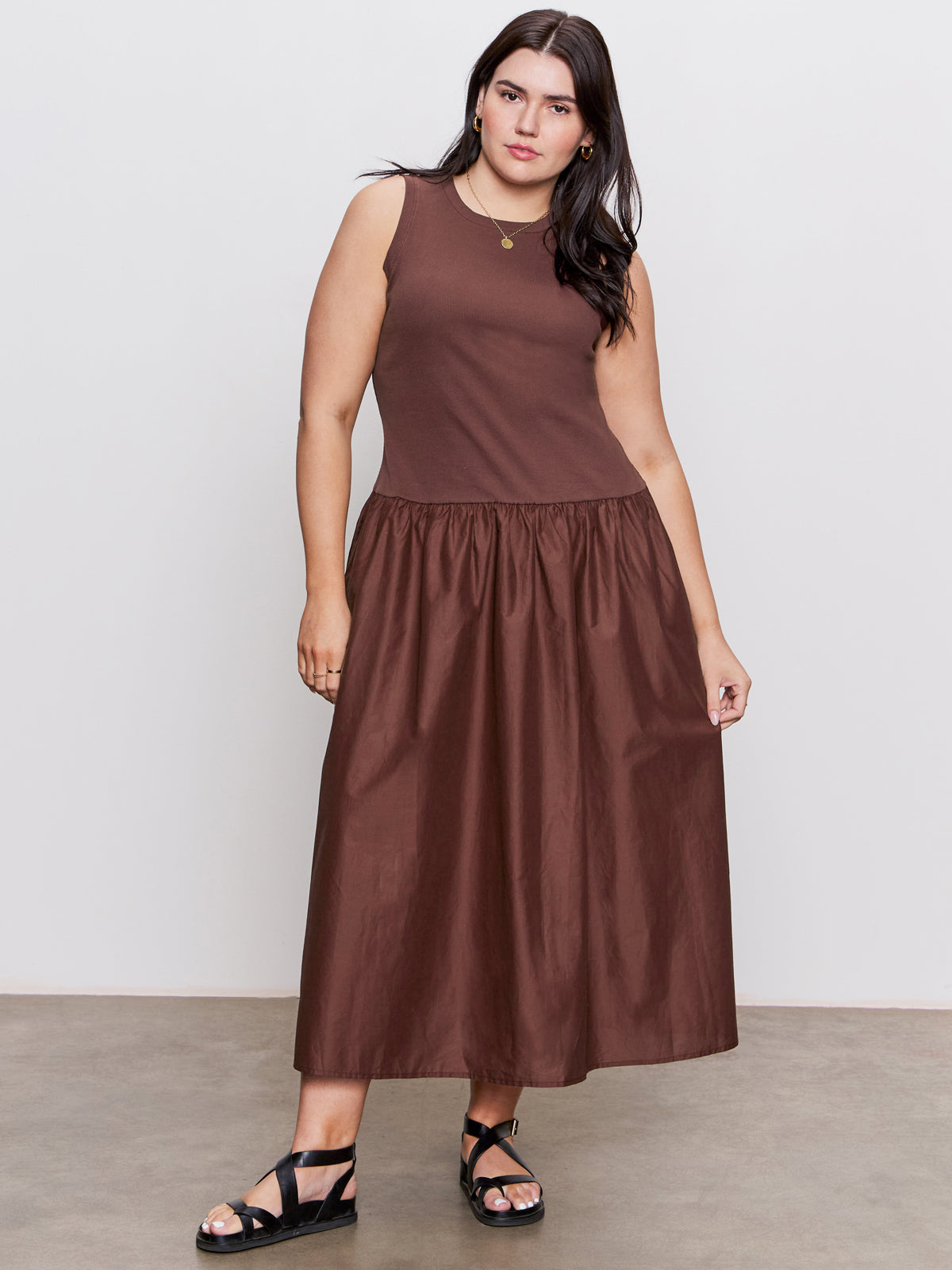 A woman with long dark hair wears Sanctuary Clothing's "take me away midi dress mesa" in extended sizing, featuring a sleeveless fitted top and gathered skirt, paired with black platform sandals. She stands on concrete before a plain light gray backdrop.