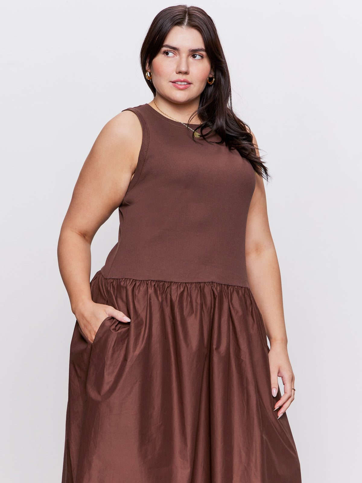 A woman with long dark hair models the Sanctuary Clothing take me away midi dress mesa in extended sizing. The sleeveless brown dress features a fitted top and gathered skirt. She stands against a plain light background, one hand in her pocket.