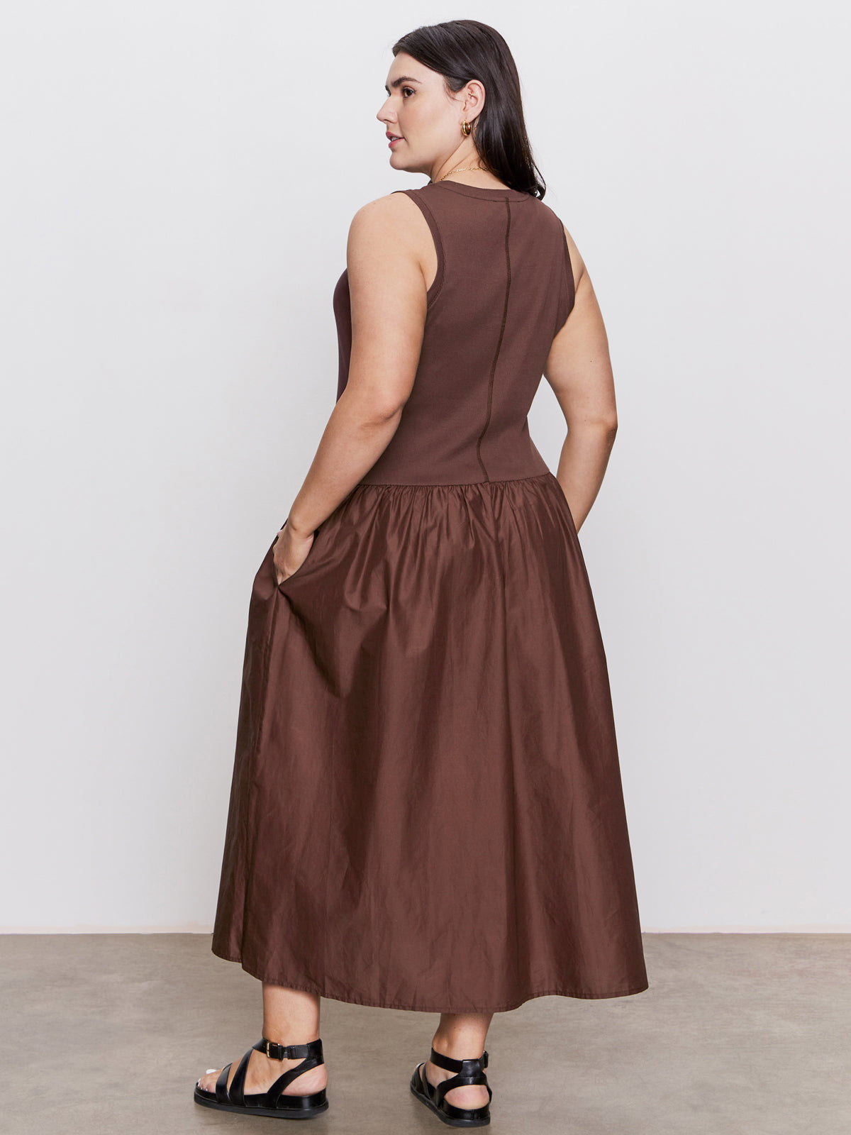 A woman faces away, wearing Sanctuary Clothing's take me away midi dress mesa (extended sizing), a sleeveless brown dress with a fitted bodice and flared skirt, paired with black sandals. She stands on a light floor against a white background.