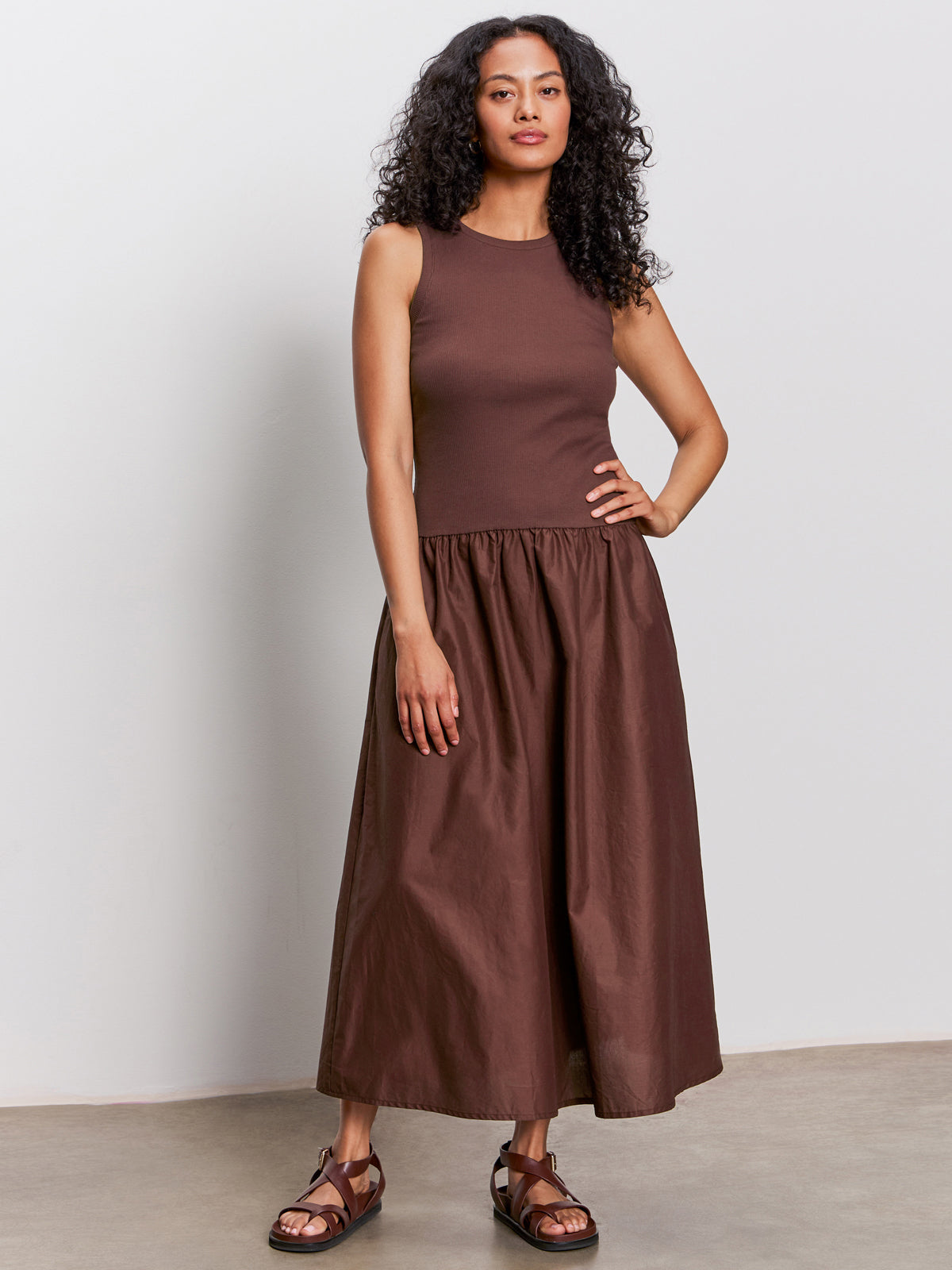 A woman with curly hair wears Sanctuary Clothing’s take me away midi dress in mesa, paired with brown sandals, standing against a plain light-colored background.