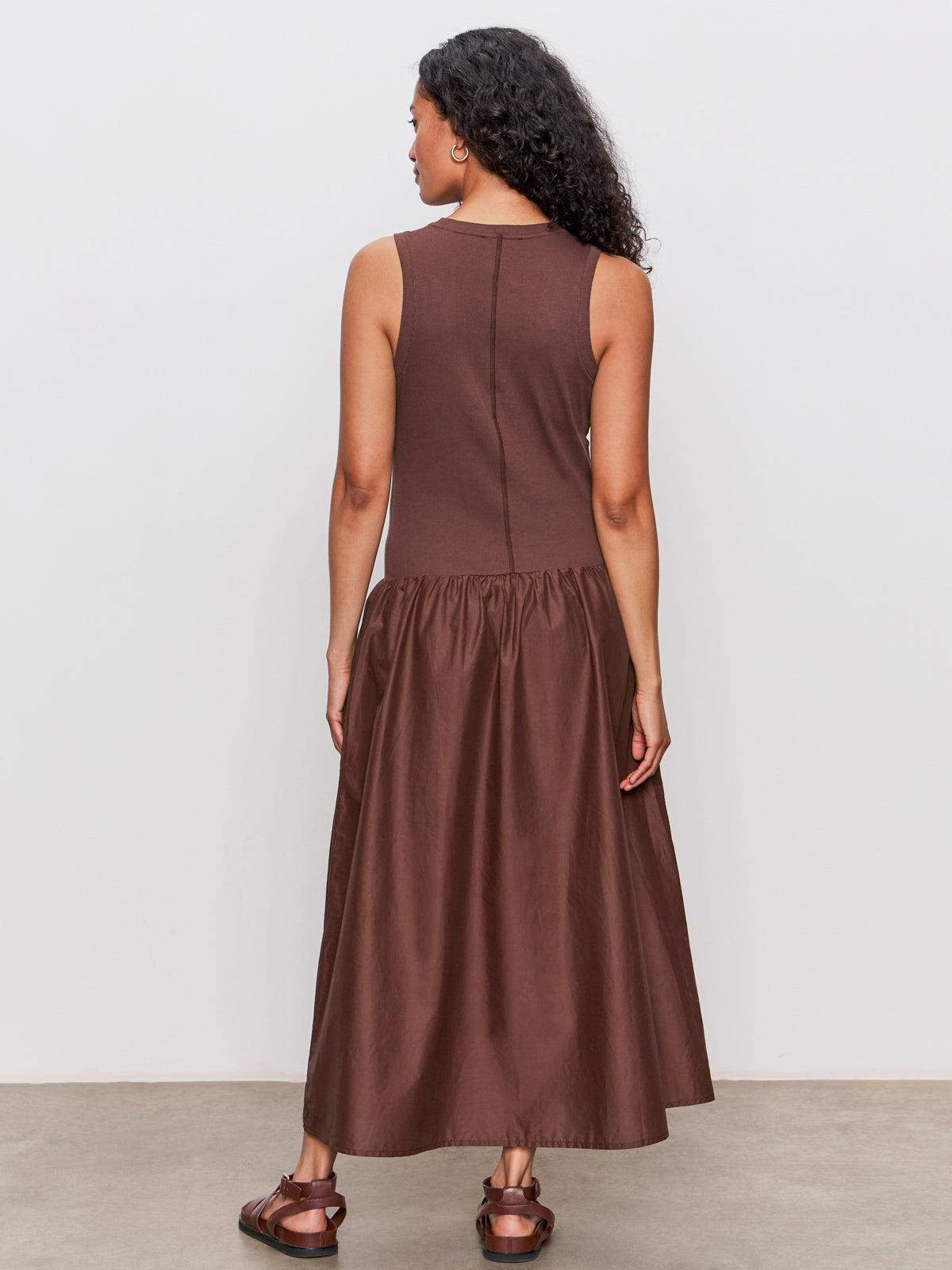 A woman with long curly hair, wearing Sanctuary Clothing's "take me away midi dress mesa," stands facing away from the camera in a sleeveless brown gathered-skirt dress and brown sandals against a plain light background.