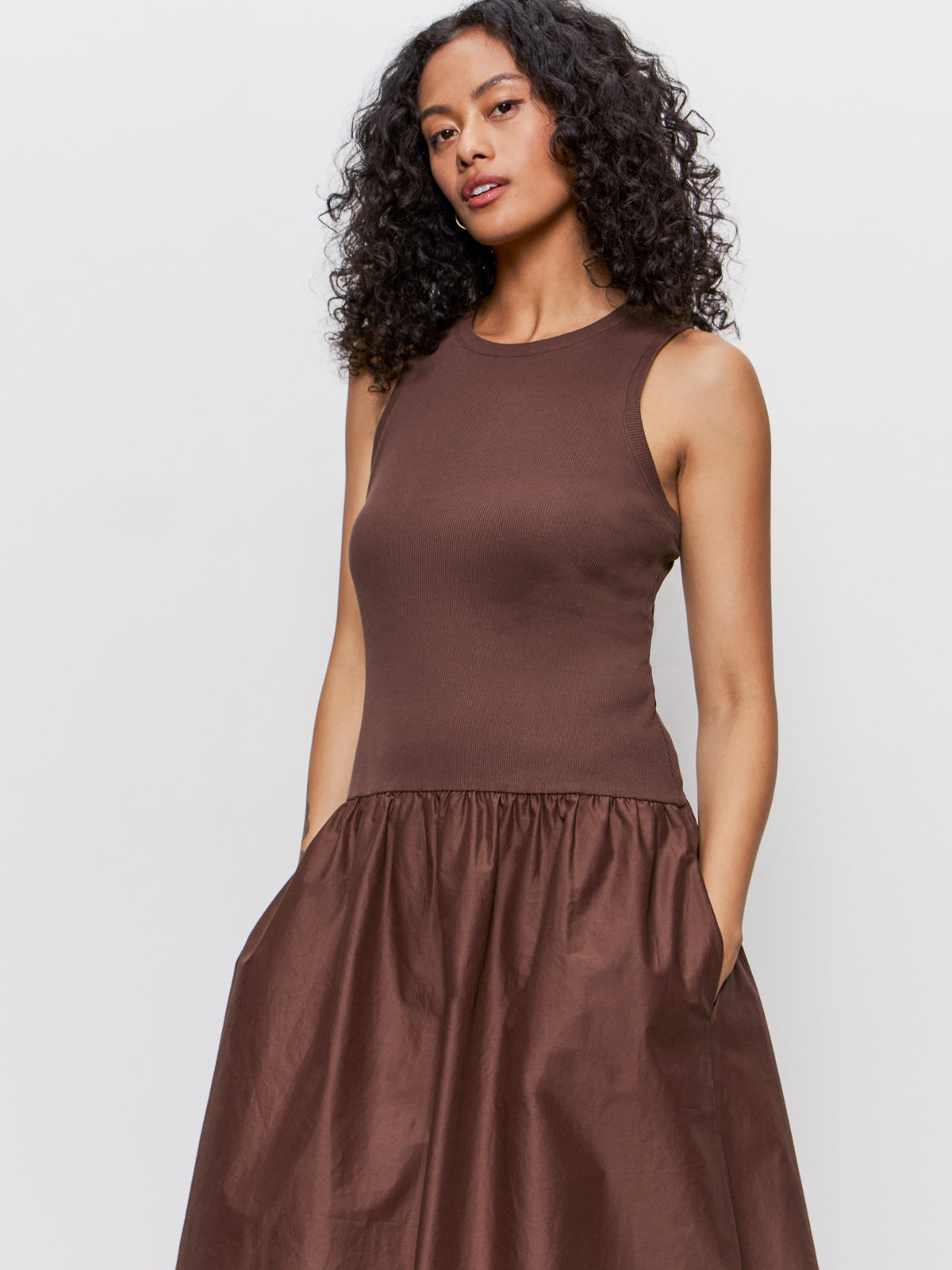 A woman with curly hair models the Sanctuary Clothing "take me away midi dress mesa"—a sleeveless, brown, fitted-top dress with a gathered skirt and pockets—standing against a plain background.