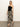 A woman stands in a minimalist room wearing the "Meet Me at Sea" black mirage midi dress by Sanctuary Clothing. Her straight, light-colored hair complements her outfit, which she finishes with black sandals.