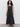 A woman with curly hair wears the Sanctuary Clothing "lucky you dress black," a sleeveless, ankle-length dress with wide straps, paired with black wedge sandals and a simple necklace against a plain light-colored wall.