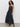 A woman with curly hair models the Sanctuary Clothing "lucky you dress black," a sleeveless, ankle-length dress, paired with black platform sandals, standing against a plain white wall.