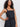A woman with curly dark hair models the Sanctuary Clothing "lucky you dress black"—a sleeveless dress with lace details—paired with a gold heart necklace, standing against a plain light background, one hand on her hip.