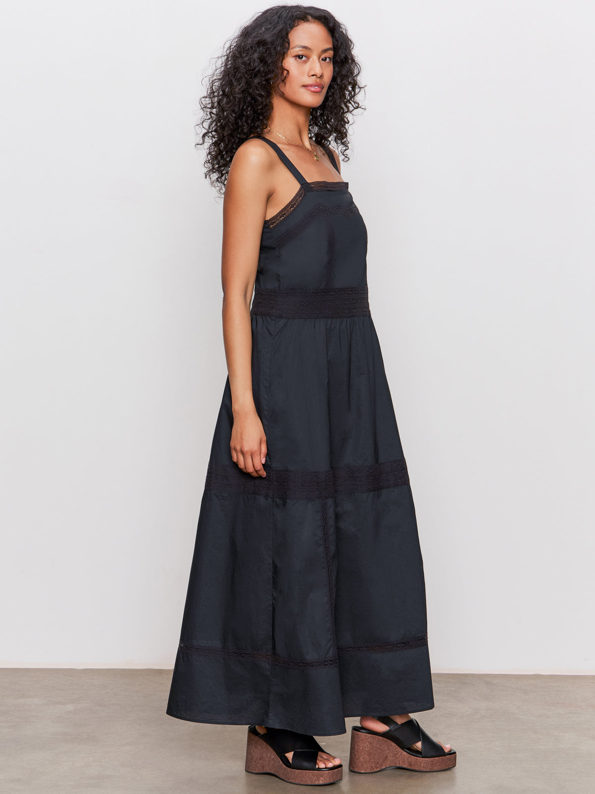 A woman with curly hair stands sideways against a white background, wearing the Sanctuary Clothing "lucky you dress black," a sleeveless, ankle-length black dress with lace details, paired with black platform sandals.