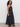 A woman with curly hair stands sideways against a white background, wearing the Sanctuary Clothing "lucky you dress black," a sleeveless, ankle-length black dress with lace details, paired with black platform sandals.