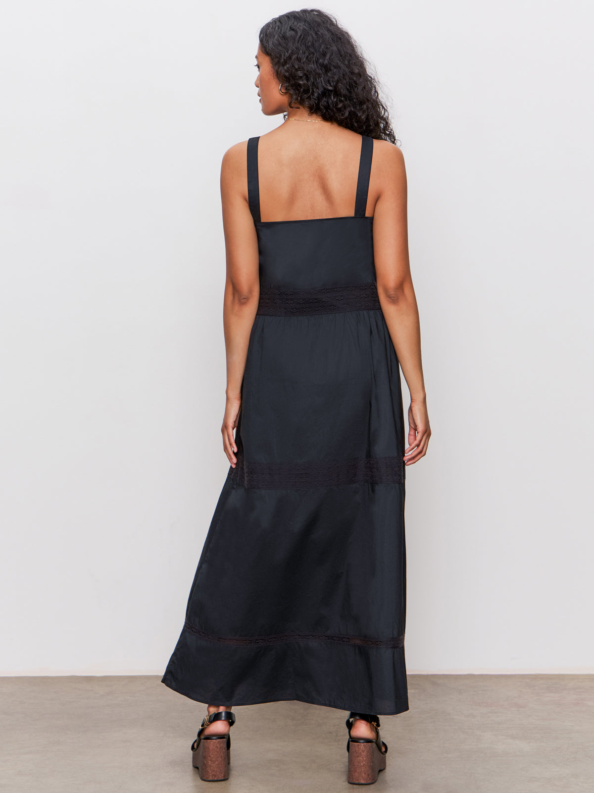 A woman with long curly hair stands facing away from the camera in Sanctuary Clothing's lucky you dress black, a sleeveless ankle-length style, paired with brown platform heels against a plain white wall and concrete floor.