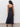 A woman with long curly hair stands facing away from the camera in Sanctuary Clothing's lucky you dress black, a sleeveless ankle-length style, paired with brown platform heels against a plain white wall and concrete floor.