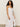 A person with long, curly hair wears a sleeveless Wilder Linen Dress in bleached white by Sanctuary Clothing. They stand against a plain beige background, gazing slightly off to the side.
