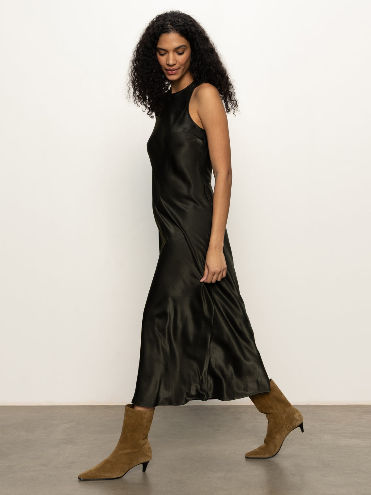 A woman with curly hair wears the Sanctuary Clothing everyday satin dress in black olive, paired with tan suede ankle boots. She walks left against a plain light background, appearing relaxed and stylish.